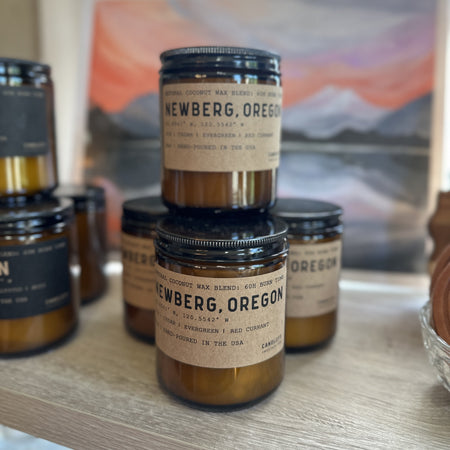 Amber glass Newberg, Oregon candle with labels on a wooden shelf, with a scenic background.