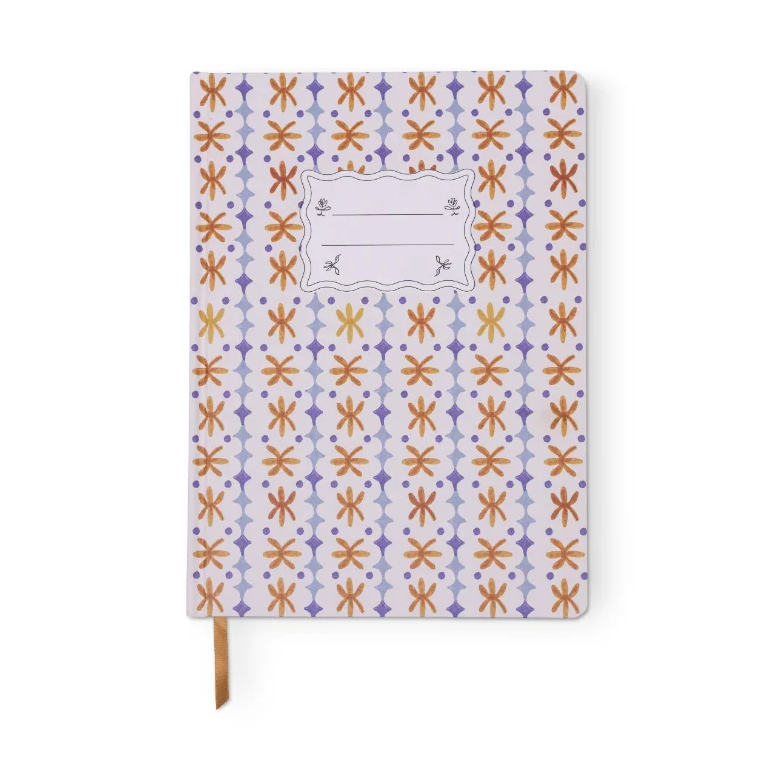 A hardcover notebook with a white cover decorated in a repeating pattern of orange and purple geometric motifs, featuring a blank decorative label in the center and a brown ribbon bookmark hanging from the bottom.