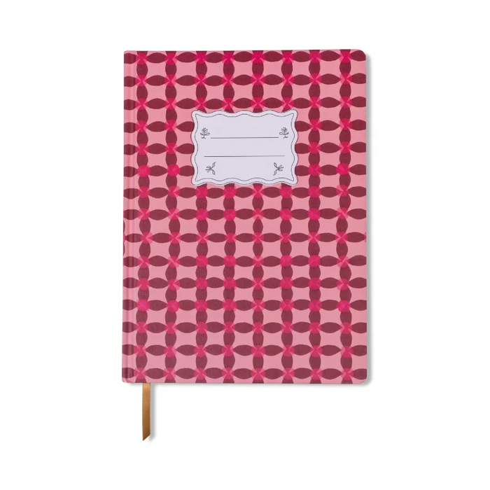 A hardcover notebook with a pink and red geometric pattern, featuring a blank white label on the front cover and a brown ribbon bookmark hanging from the bottom edge, set against a plain white background.