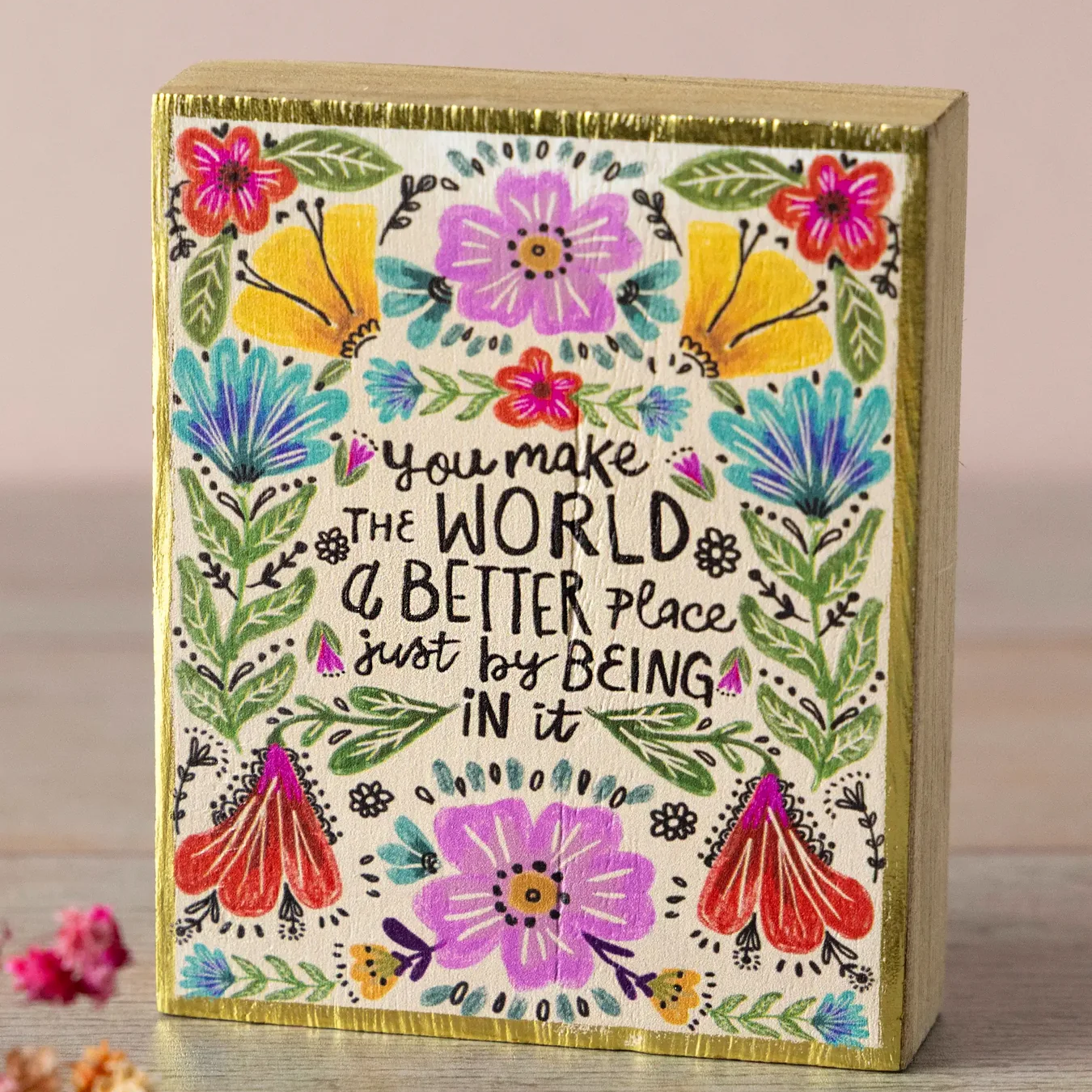 A rectangular decorative block with a gold-toned border and a cream front panel covered in colorful floral illustrations and the handwritten quote, 'you make THE WORLD a BETTER place just by BEING IN it.' The block sits on a wooden surface against a soft beige background, with a few small dried flowers and coins nearby.