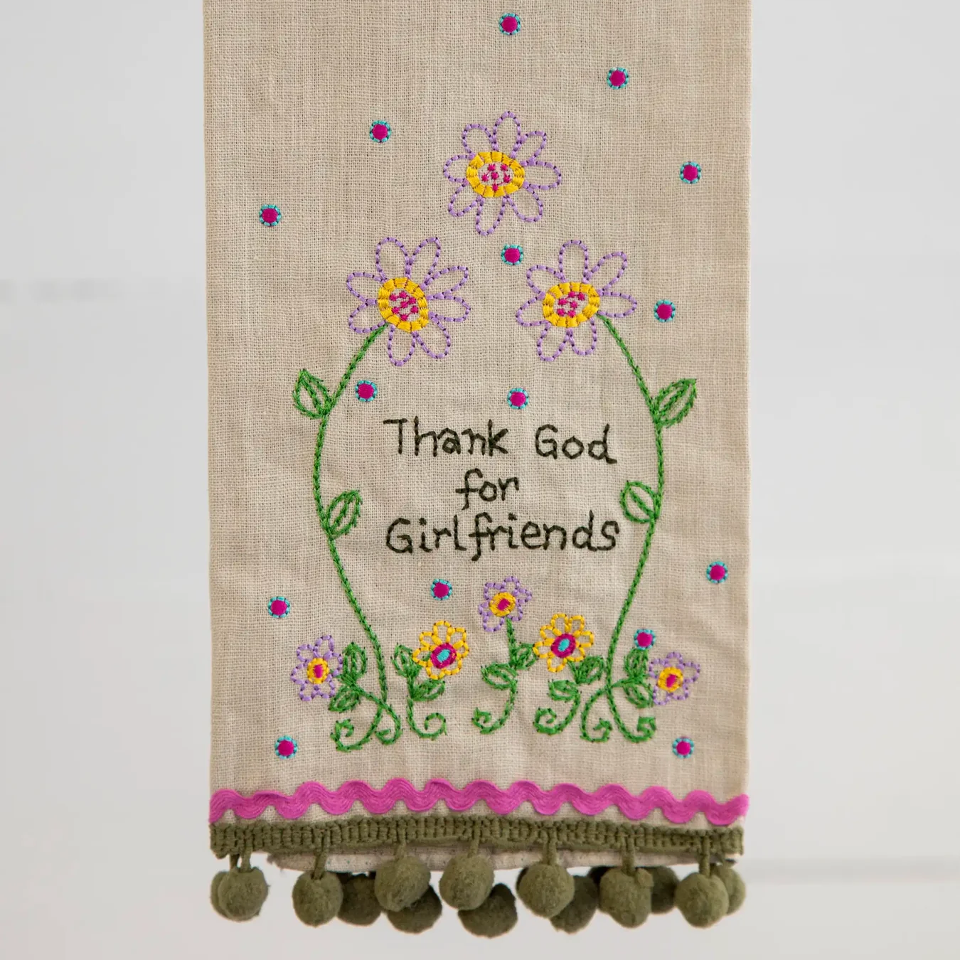 A beige linen tea towel with embroidered flowers and the text 'Thank God for Girlfriend' hanging from a rustic wooden dowel. The bottom of the towel features a pink embroidered scalloped border and a row of green pom-poms.
