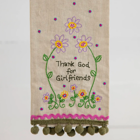 A beige linen tea towel with embroidered flowers and the text 'Thank God for Girlfriend' hanging from a rustic wooden dowel. The bottom of the towel features a pink embroidered scalloped border and a row of green pom-poms.