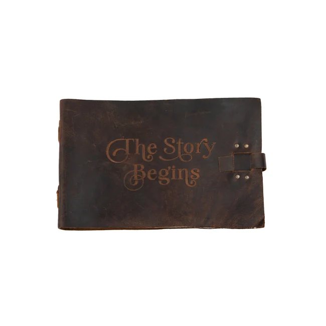 A dark brown leather journal with 'The Story Begins' embossed on the front cover, featuring a strap closure on the right side.