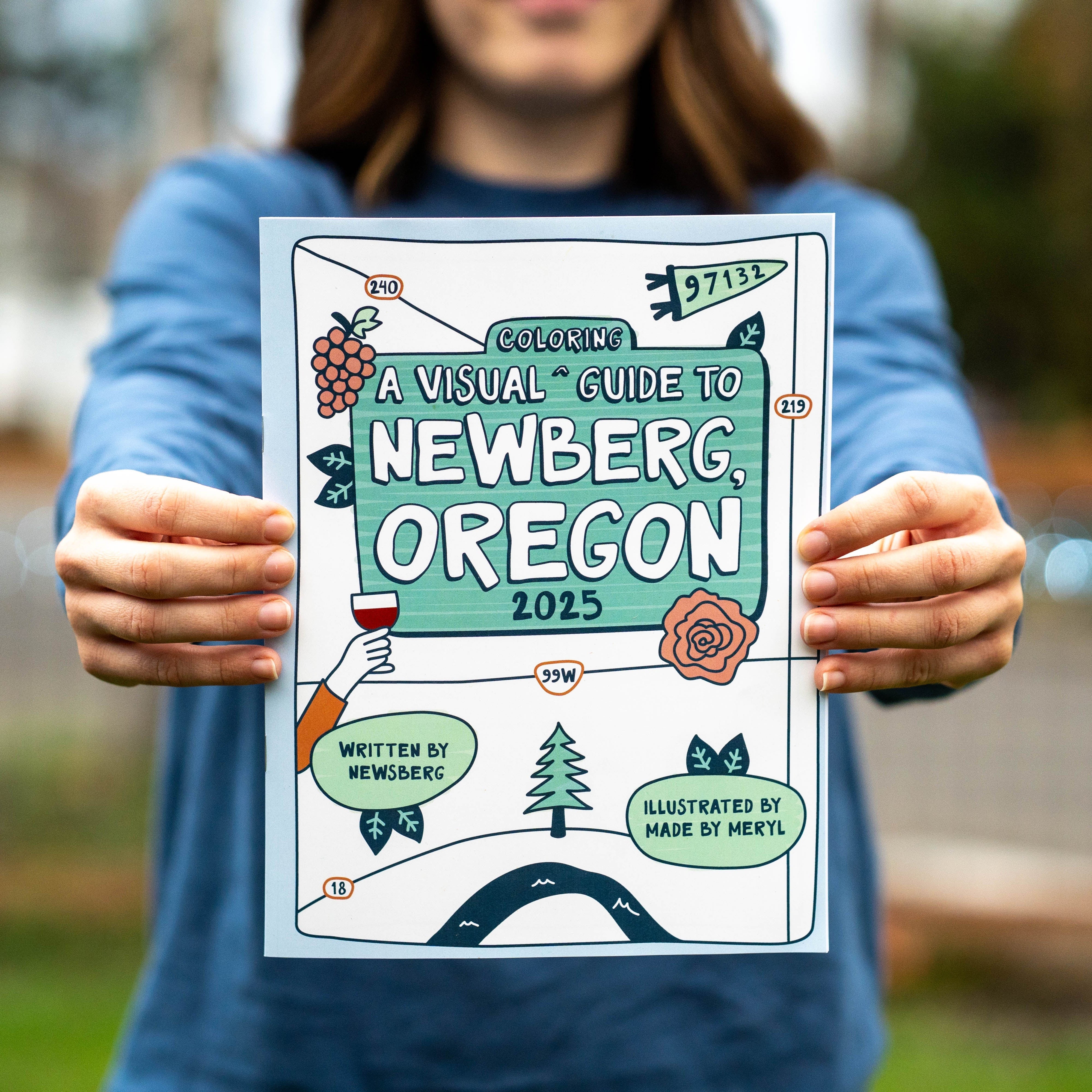 Person holding a colorful coloring book titled 'A Visual Guide to Newberg, Oregon 2025' outdoors.