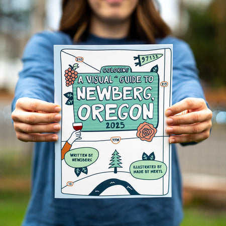 Person holding a colorful coloring book titled 'A Visual Guide to Newberg, Oregon 2025' outdoors.