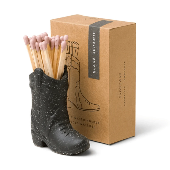 Black ceramic boot-shaped match holder with matches next to a cardboard box on a white background