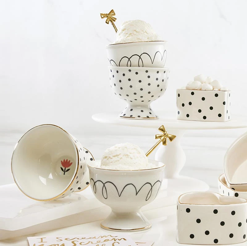 A set of white ceramic dessert bowls and cups with black polka dots, black wavy line patterns, gold trim, and small floral details, styled with ice cream on a white background.
