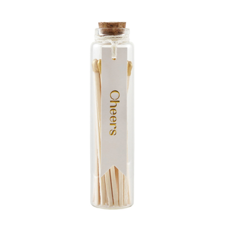 A clear glass bottle with a cork stopper, filled with wooden matchsticks and wrapped with a white paper tag that reads 'Cheers' in gold lettering, on a white background.