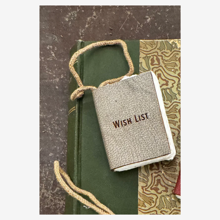 Small book ornament labeled 'Wish List' tied with string on a textured surface.