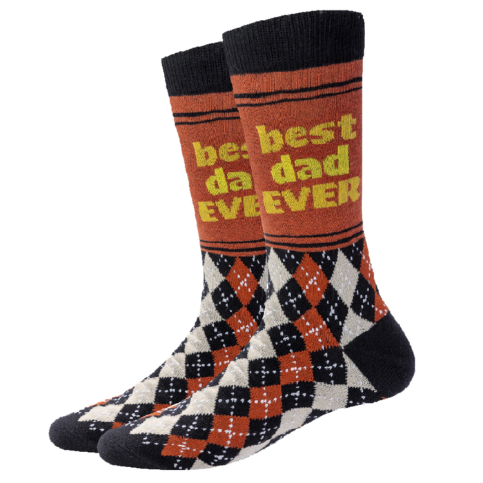 A pair of crew socks with a black cuff and toe, featuring a black, orange, and white argyle pattern on the lower half and a rust-orange upper half with yellow text that reads 'best dad EVER'.