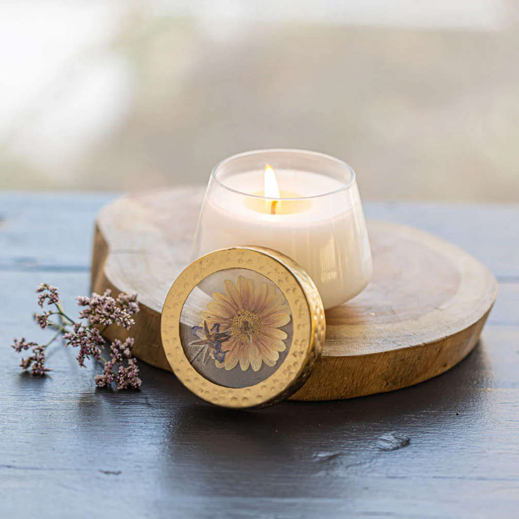 Candle in a glass holder with a wooden lid featuring a pressed flower design, on a wooden surface.