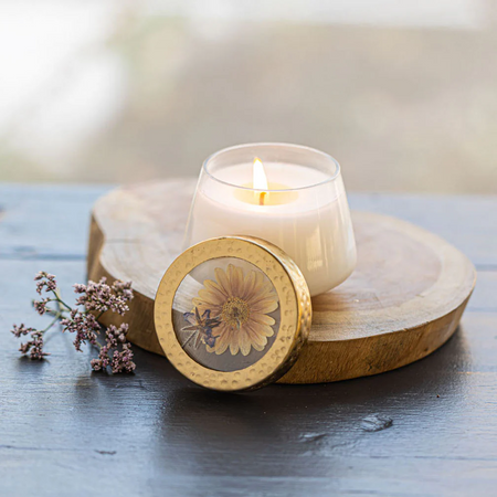 Candle in a glass holder with a wooden lid featuring a pressed flower design, on a wooden surface.