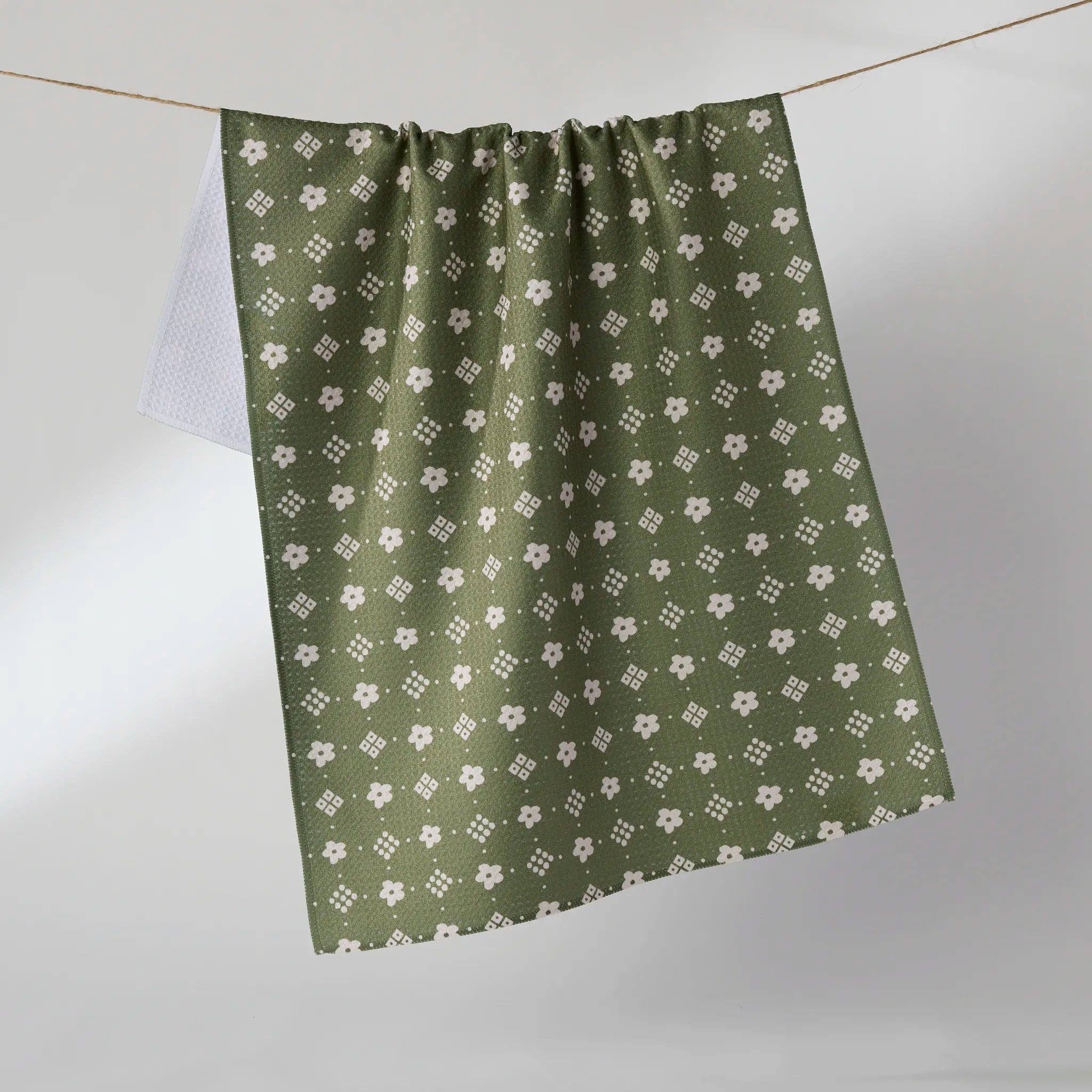 An olive green towel with a repeating white floral and geometric pattern, hanging from a thin wire against a neutral gray background.