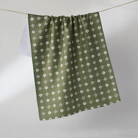 An olive green towel with a repeating white floral and geometric pattern, hanging from a thin wire against a neutral gray background.