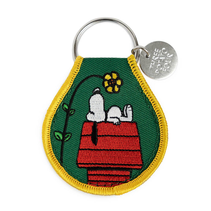 Keychain with Snoopy and a flower on a doghouse design, featuring a green background and yellow border.