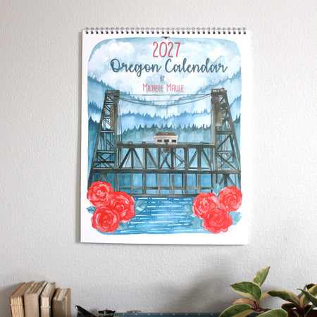 A 2027 Oregon Calendar by Michele Maule featuring a watercolor illustration of a bridge over blue water with red roses, hanging on a light gray wall 