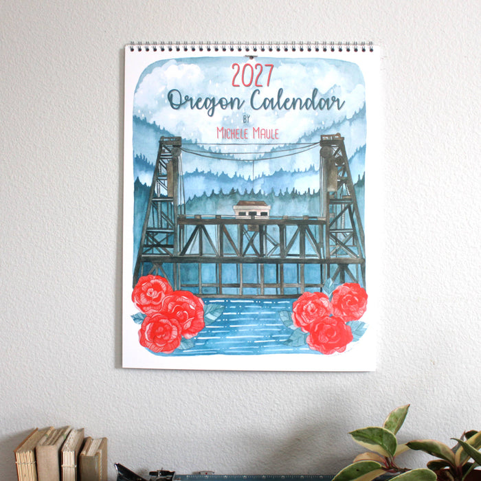 A 2027 Oregon Calendar by Michele Maule featuring a watercolor illustration of a bridge over blue water with red roses, hanging on a light gray wall 