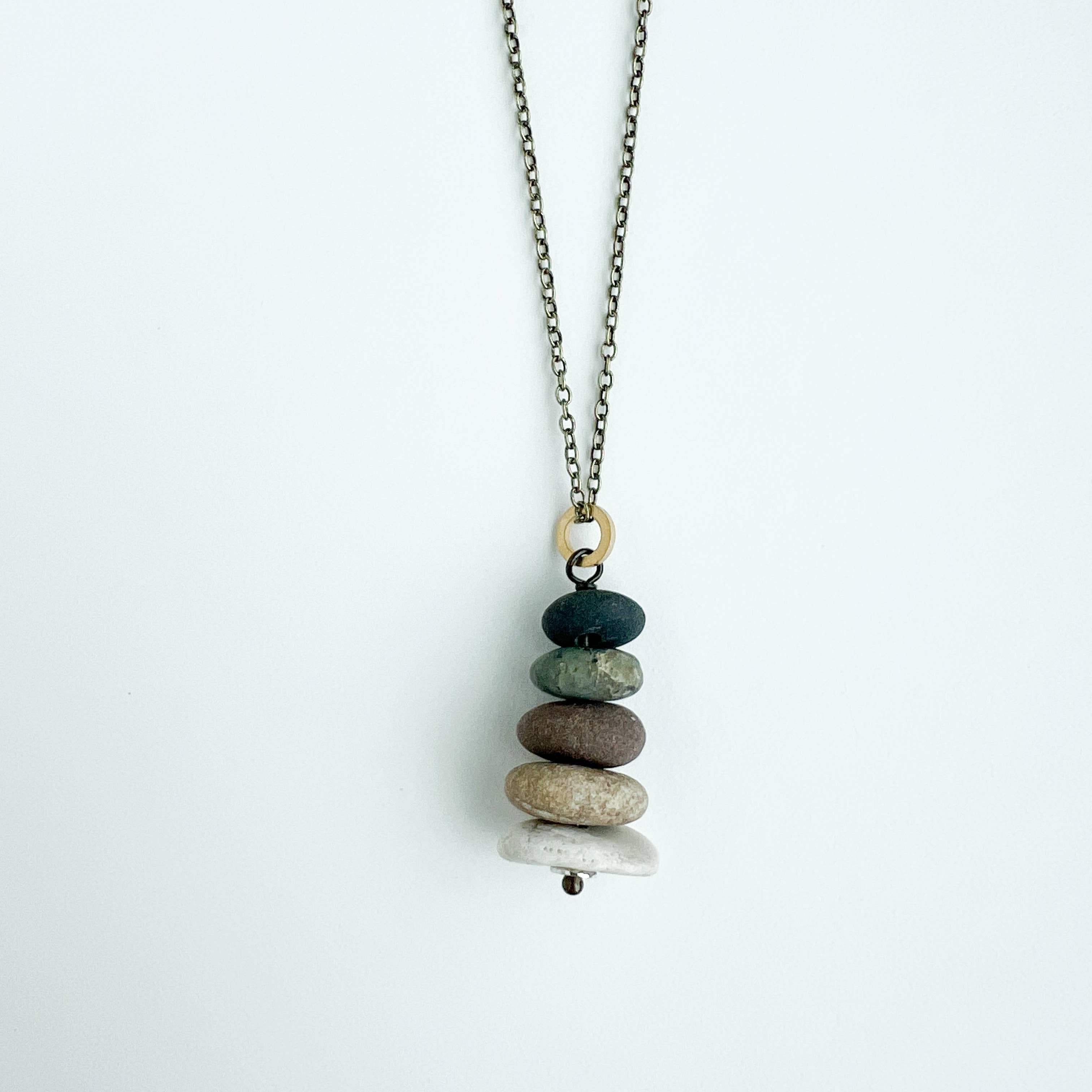 A delicate necklace with a stacked pebble pendant on a light gray background. The pendant features a stack of seven smooth stones in shades of dark green, brown, beige, and white, suspended from an antique bronze chain.
