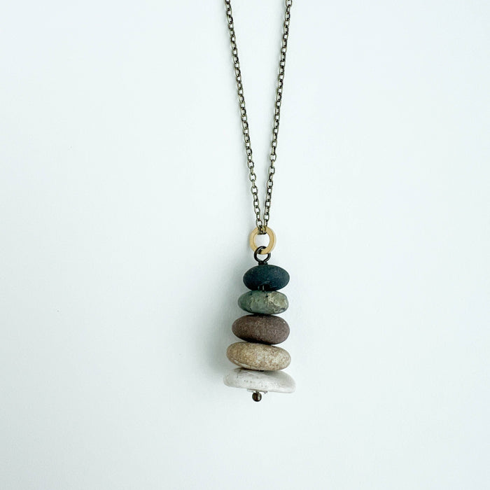 A delicate necklace with a stacked pebble pendant on a light gray background. The pendant features a stack of seven smooth stones in shades of dark green, brown, beige, and white, suspended from an antique bronze chain.