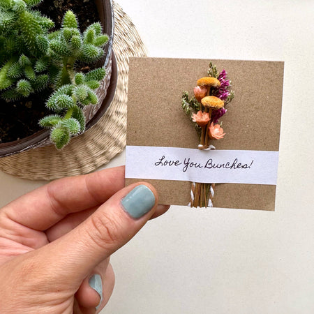 Hand holding a card with dried flowers and 'Love You Bunches!' text, next to a potted plant.