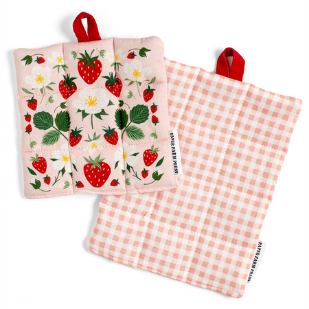 Two quilted fabric potholders from Sweet Farm Fools, displayed on a clean white background. One potholder shows a pink background with a strawberry and white flower pattern, complete with a red hanging loop.
