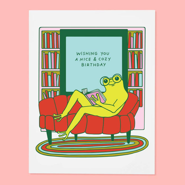 Frog on a red couch with book, text 'Wishing you a nice & cozy birthday' in a room with bookshelves.