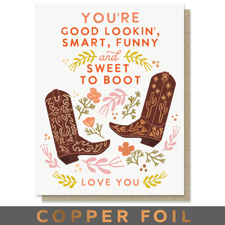 A greeting card with a western theme, featuring the text 'YOU'RE GOOD LOOKIN', 'SMART, FUNNY and SWEET TO BOOT' and 'LOVE YOU' in orange lettering, decorated with illustrated brown cowboy boots, flowers, and leaves. A kraft envelope is visible behind the card, and a dark banner at the bottom reads 'COPPER FOIL'.