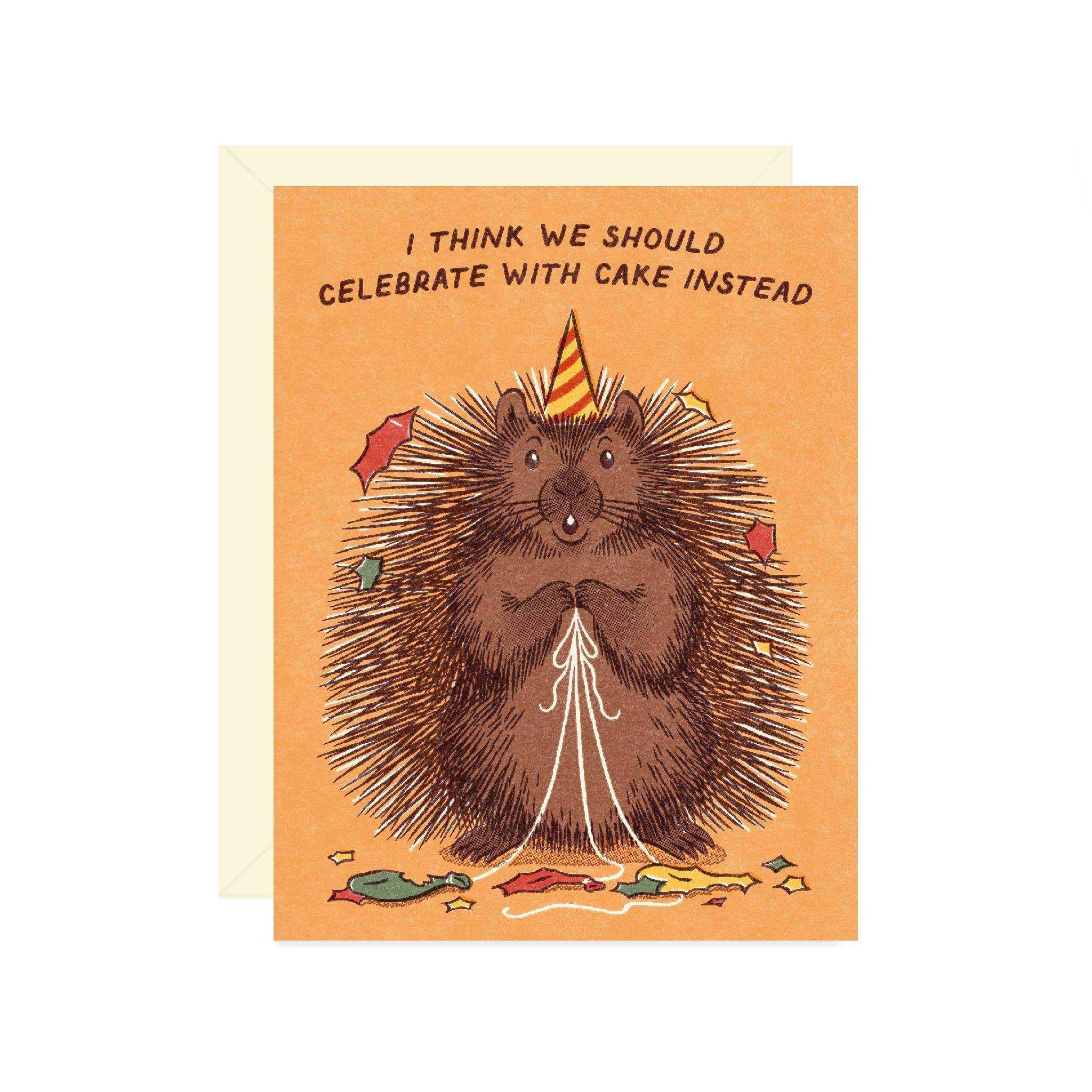 A greeting card featuring a brown hedgehog wearing a party hat and holding popped balloons, with the text 'I THINK WE SHOULD CELEBRATE WITH CAKE INSTEAD' above it. The card has an orange background and is shown with a cream envelope behind it on a white background.