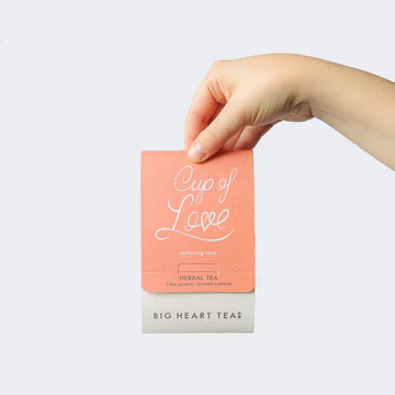 Hand holding a pink tea box labeled 'Cup of Love' by Big Heart Tea on a light gray background