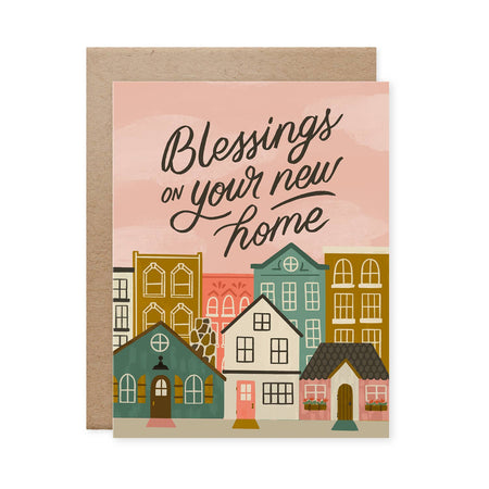 A housewarming card with a hand lettered message 'Blessings on your new home' accompanied by an illustration of three houses in different colors. The card is shown with a kraft envelope.