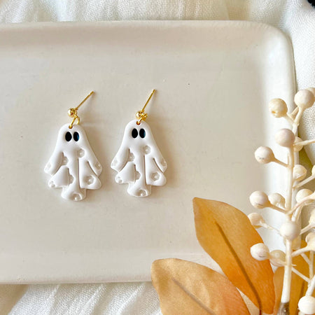 A pair of white ghost-shaped dangle earrings with gold hooks, displayed on a white rectangular tray with styled white fabric, a black-and-white patterned textile, and dried beige leaves and berries.