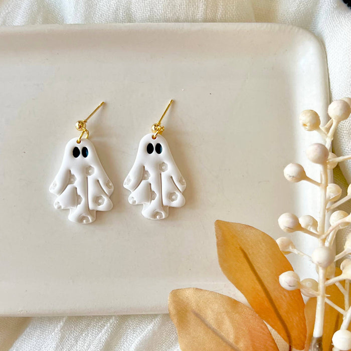 A pair of white ghost-shaped dangle earrings with gold hooks, displayed on a white rectangular tray with styled white fabric, a black-and-white patterned textile, and dried beige leaves and berries.