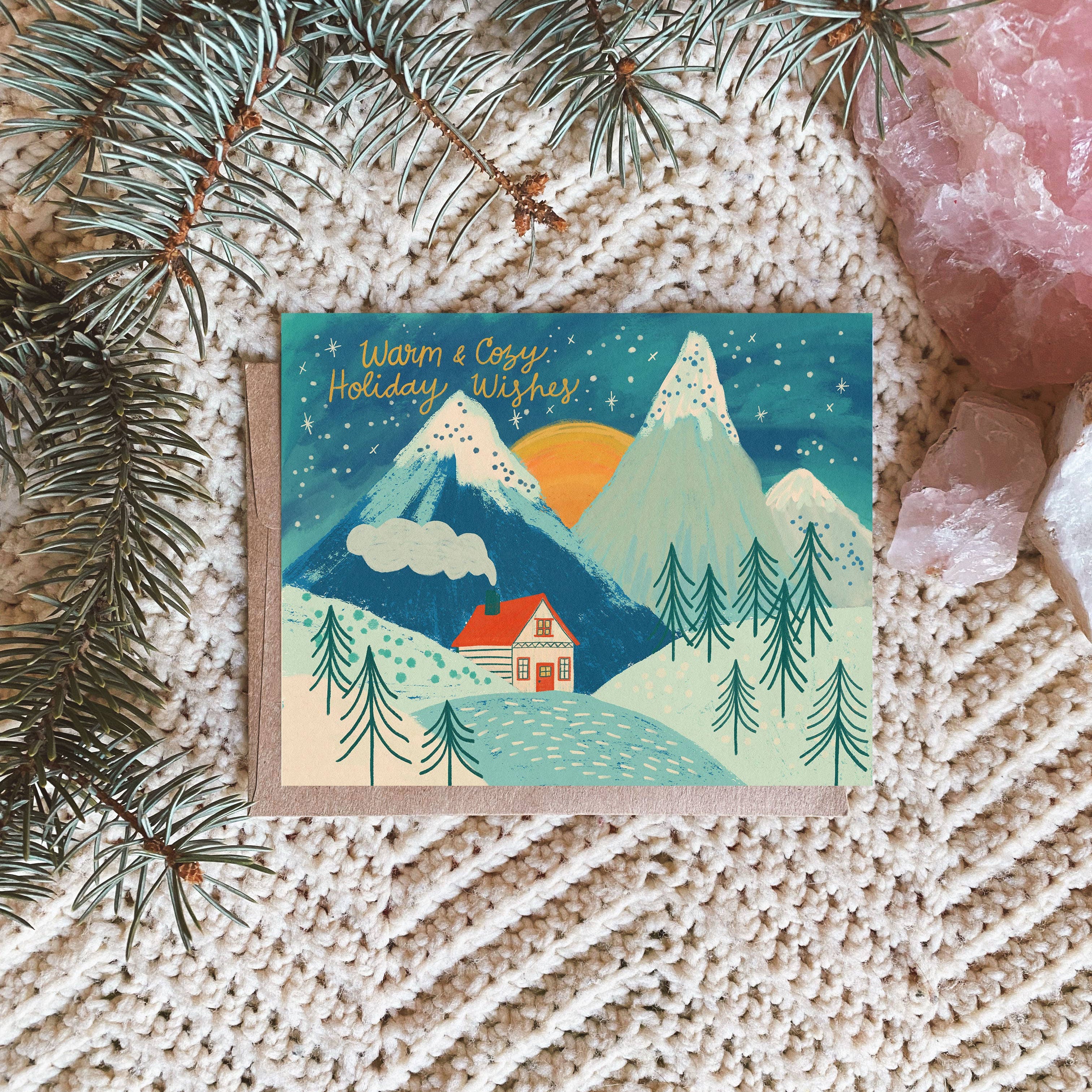 A holiday greeting card with an illustration of a cabin in a snowy landscape, under a blue sky with stars, placed on a textured fabric backdrop with pine branches and crystals.
