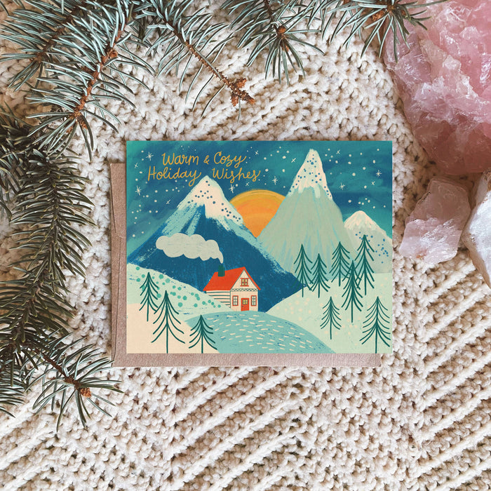 A holiday greeting card with an illustration of a cabin in a snowy landscape, under a blue sky with stars, placed on a textured fabric backdrop with pine branches and crystals.