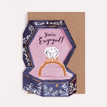A die cut greeting card featuring an illustrated open hexagonal ring box with a diamond ring inside and the words 'You're Engaged!' on a pink interior, set against a kraft brown card on  a white background.
