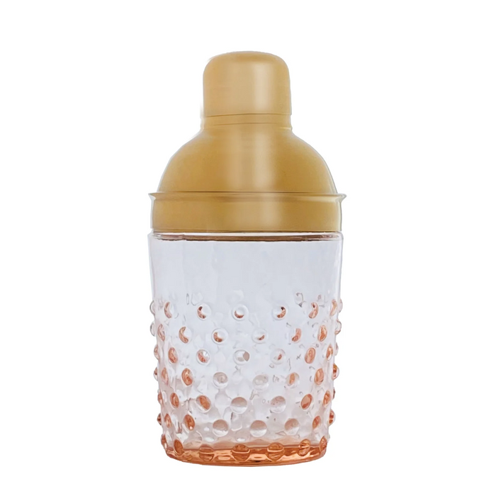 A cocktail shaker with a translucent clear glass body featuring a textured bubble pattern that fades into a warm peach-orange at the bottom, topped with a metallic gold screw-on lid.