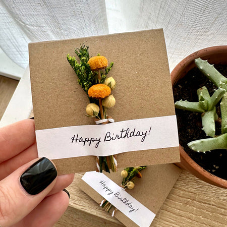 Hand holding a 'Happy Birthday!' card with floral design and potted plant in the background
