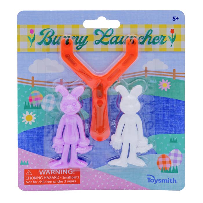 The product image shows a Bunny Launcher toy consisting of an orange slingshot and two rubber bunny figures in pink and white colors, packaged on a cardboard backing with a springtime theme illustration.