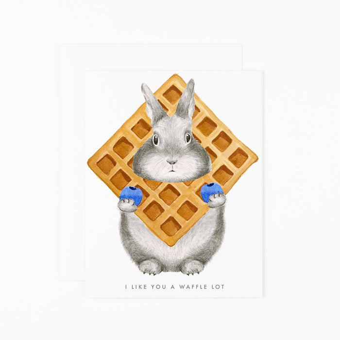 A greeting card with an illustration of a bunny peeking through a waffle grid, with the text 'I like you a waffle lot' on the front. The card is paired with a matching soft white envelope.