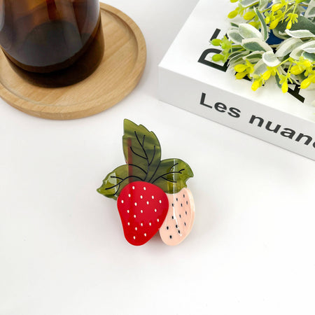Strawberry-shaped hair clip on a white surface with a book and plant in the background