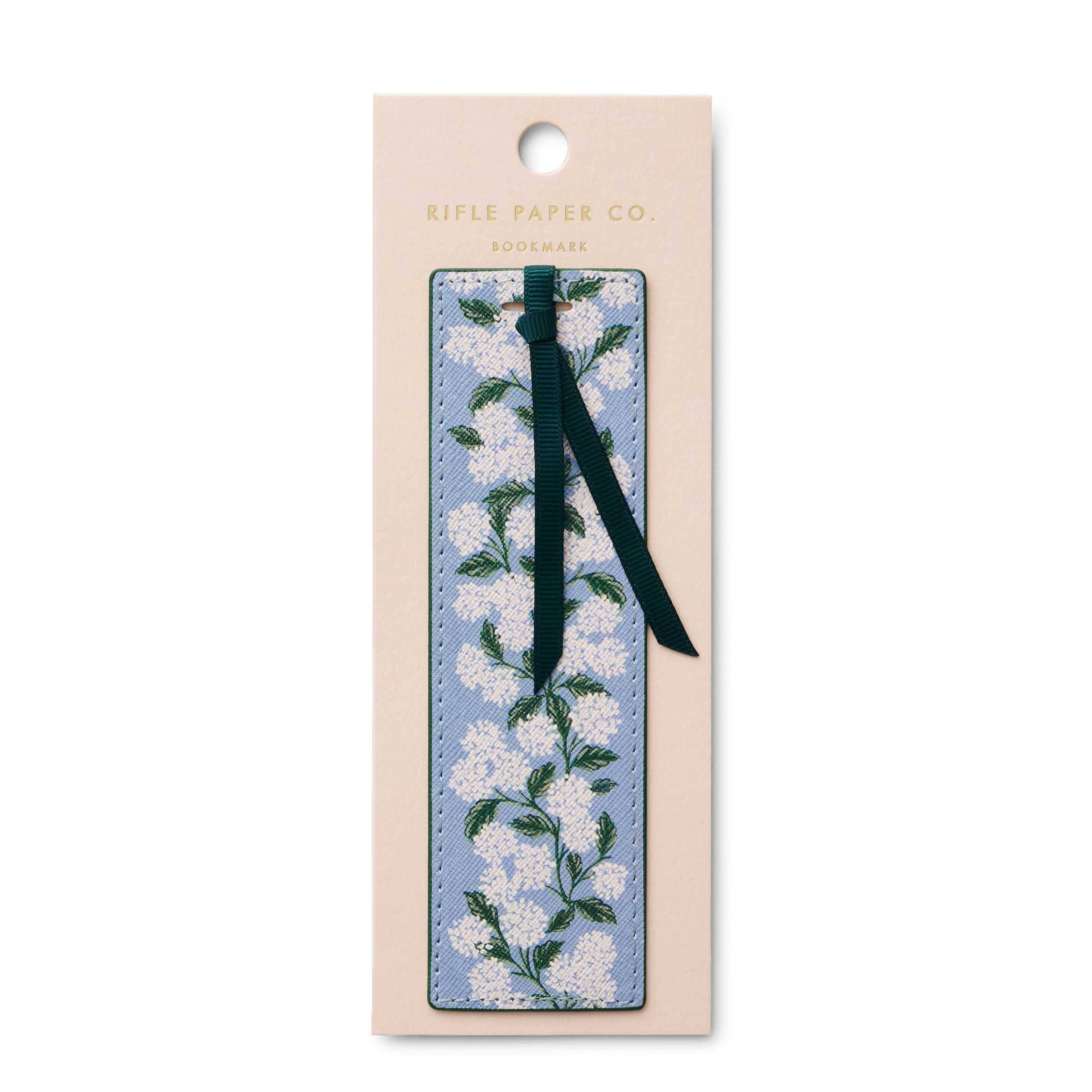 A floral bookmark by Rifle Paper Co. i, featuring a light blue background with white blossoms and green leaves, attached to a dark green ribbon and displayed on its beige backing card against a white background.