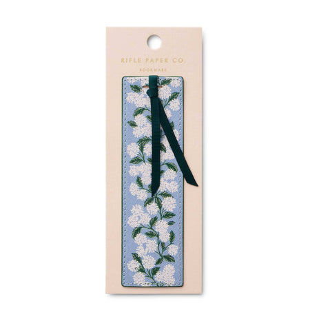 A floral bookmark by Rifle Paper Co. i, featuring a light blue background with white blossoms and green leaves, attached to a dark green ribbon and displayed on its beige backing card against a white background.