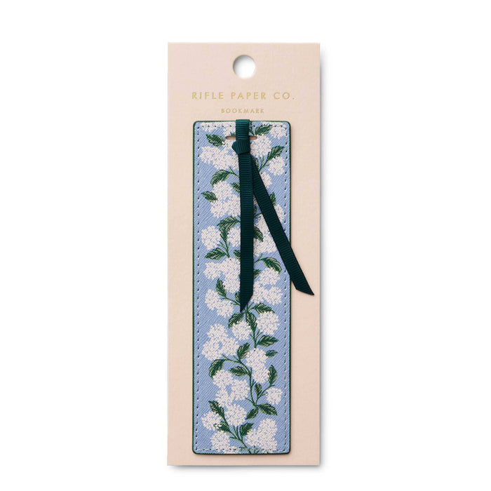 A floral bookmark by Rifle Paper Co. i, featuring a light blue background with white blossoms and green leaves, attached to a dark green ribbon and displayed on its beige backing card against a white background.