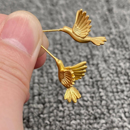 Gold bird-shaped earrings held between fingers against a textured gray background