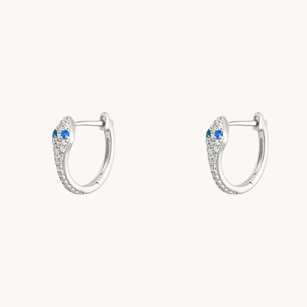 Silver snake huggie earrings with blue sapphire eyes and sparkling accents on a white background.