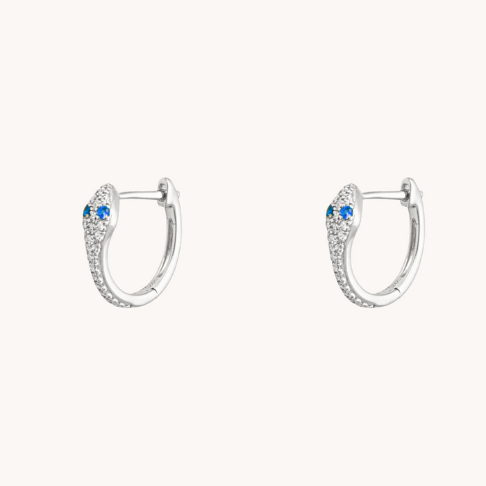 Silver snake huggie earrings with blue sapphire eyes and sparkling accents on a white background.