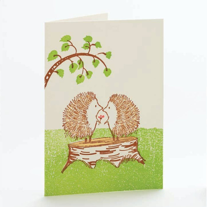 A notecard with a hand-drawn illustration of two hedgehogs on a tree stump, with a white envelope.